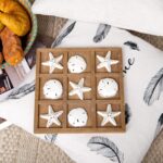 A distressed white NIKKY HOME Beach Tic Tac Toe game board with starfish and sand dollar resin pieces.